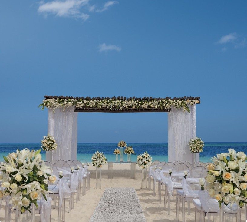 A beach wedding ceremony with a white draped gazebo, flowers, and chairs facing the turquoise Caribbean sea